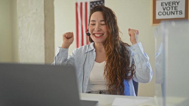Woman with headset and laptop raises fists in celebration inside a voting building with american flag and 'every vote counts' sign; civic pride joy.