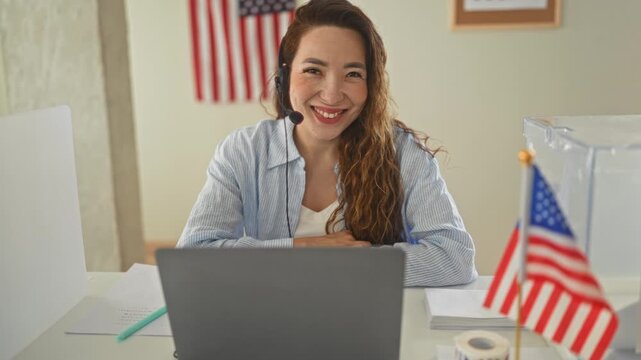 Woman at laptop wearing headset smiles beside american flag and clear ballot box on a desk in a polling building; civic pride.