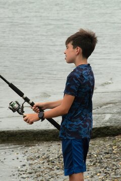 A young boy stands on a pebbled shore concentrating as he reels in a line at a calm, overcast riverbank, capturing quiet patience, outdoor recreation and a summertime hobby by the water