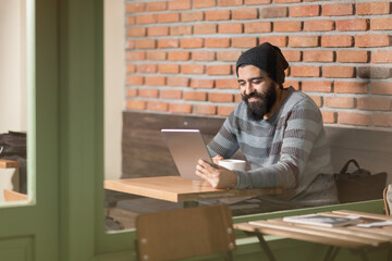 Side view of a focused bearded man wearing a beanie using a digital tablet while sitting at a table in a modern cafe with a brick wall background. © Fatih