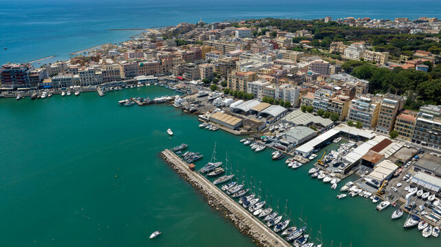 Aerial view of the coastal town of Anzio, near Rome in Italy, showing the bustling port with boats and yachts, turquoise sea waters, breakwaters, and colorful urban buildings along the coastline.