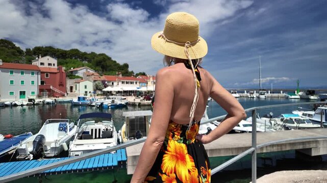Woman enjoying a summer day, walking on a vibrant boardwalk in Luka Rovenska harbor surrounded by colorful traditional houses and boats on Losinj Island, Croatia