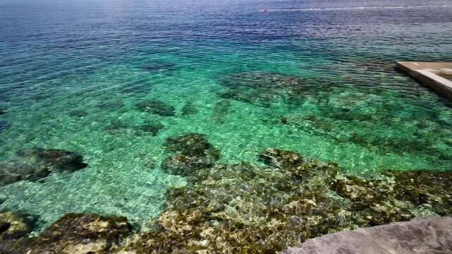 Clear turquoise water washing over natural rock pools and concrete seawalls along the waterfront harbor in Luka Rovenska, Losinj Island, Croatia, offering a refreshing summer swimming experience