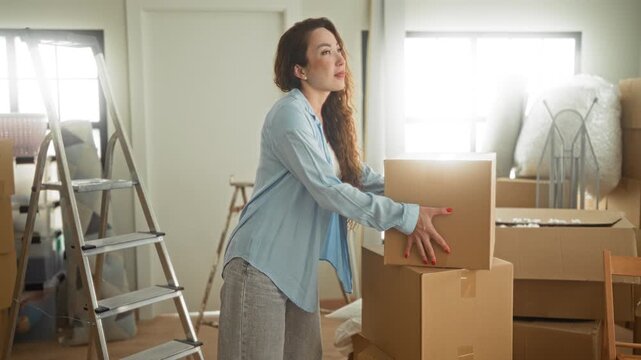 Woman lifting a box and looking up amid packed cartons and a stepladder inside a residential building, placing packages on a stack; hopeful new beginning.