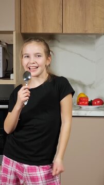 Cheerful young girl in pajamas having fun in the kitchen, dancing energetically and singing into a spoon as if it were a microphone, embodying pure joy and playful childhood