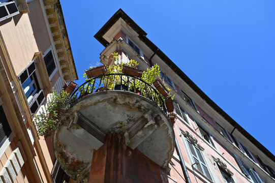 classic old circular metal balcony. black steel picket balustrade and railing. flowers in plastic pots. deteriorating cantilevered concrete slab. spalling stucco exterior elevation. decorative window.
