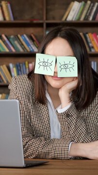 Young businesswoman with sticky notes featuring drawn eyes on her face, simulating wakefulness while feeling bored and tired at her workplace desk with a laptop in a library