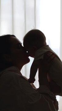 Silhouette of a loving mother tenderly holding her adorable newborn baby, lifting the infant in the air and sharing a gentle kiss against a brightly lit window in a warm, cozy room