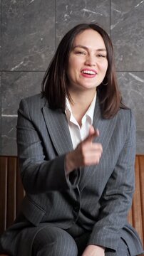 Cheerful female entrepreneur in a stylish business suit looking directly at the camera, actively gesturing, waving hello, making a heart shape, and showing an okay sign with a smile