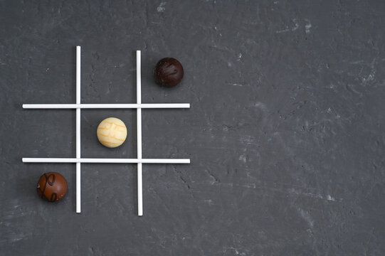 Chocolate candies arranged as tic tac toe game on dark stone background with white grid lines. Chocolate balls act as players pieces creating playful strategy mood. concept for fun dessert, game idea