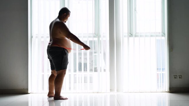 Overweight Man Measuring Waist with Red Tape Measure Standing Near Window