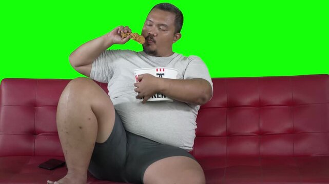 Overweight Asian Man Eating Fried Chicken Bucket on Red Sofa Isolated Green Screen