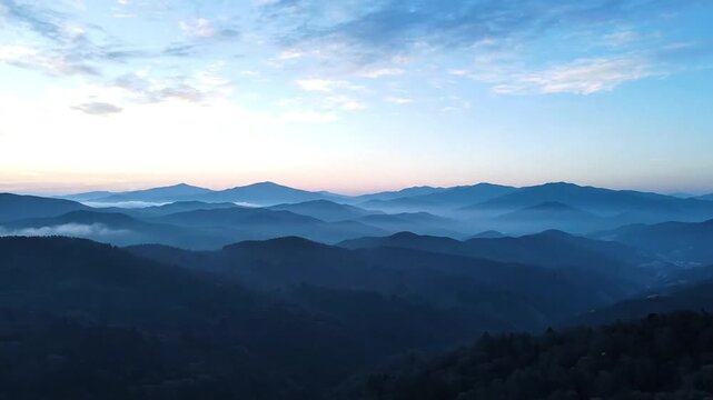 Layers of distant mountain ridges gradually appearing as morning fog slowly clears under soft blue sunrise light. Slow atmospheric movement with subtle cloud drifting creating emotional tranquility. 