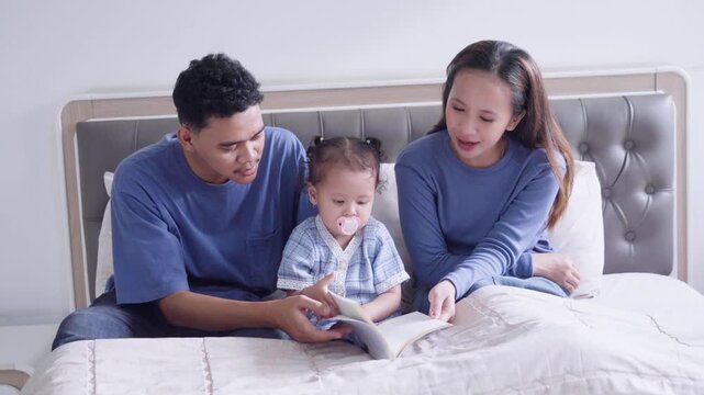 Happy Asian Parents Reading Book with Cute Toddler Daughter on Bed