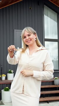 Happy female real estate agent holding keys while standing in front of a modern house, a successful businesswoman celebrating the sale of a new property and showing the keys to the camera
