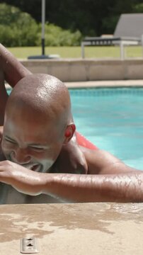 Vertical video: Laughing dad in trunks holding pool as son climbing on back in red trunks playing