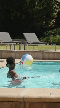 Vertical video: Chasing youth in red trunks kicking drifting beach ball in home pool, adult joining