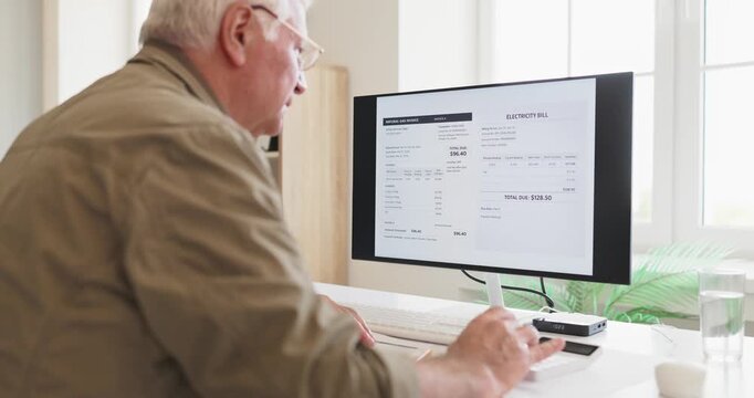 Senior elderly man checks electricity bill on computer. At a home desk retiree reviews utilities invoice, calculates costs, and prepares online payment. Clear concept of energy cost control.