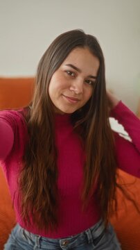 Woman touching long hair with hand while taking a selfie on bed in bedroom, wearing pink sweater and jeans and seated on orange bedspread, smiling at camera; serene confidence.