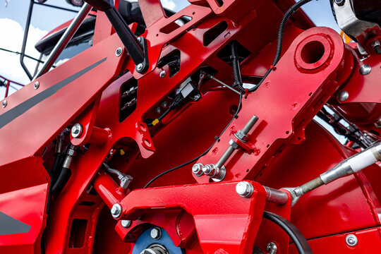 Agricultural machinery linkage adjustment mechanism section. Modern farm equipment crop harvesting assembly on red metal frame with hydraulic hoses and industrial vehicle technical details.