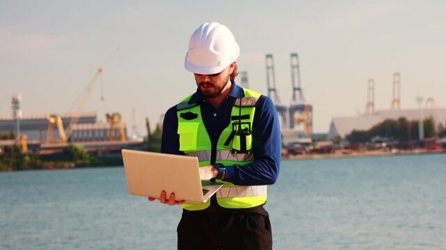 Male architect engineer carrying laptop works outdoors inspecting port construction site wearing safety uniform including safety glasses and helmet demonstrating strong awareness safety  in the field.