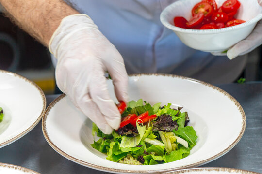 Close up of a professional chef hand arranging fresh green salad with cherry tomatoes on a plate in a restaurant kitchen.
