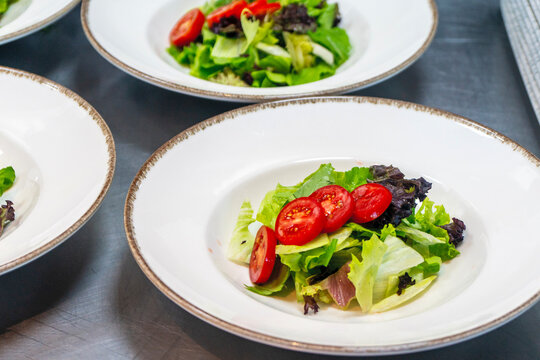 Close up of a professional chef hand arranging fresh green salad with cherry tomatoes on a plate in a restaurant kitchen.