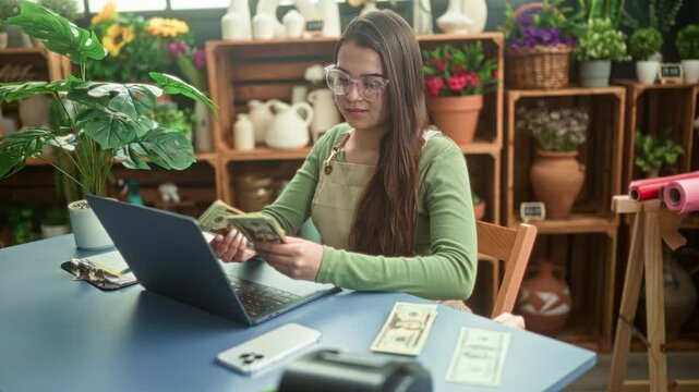 Woman florist counts cash and holds bills in hand while working at studio desk with laptop and flowers on shelves; small business focused.