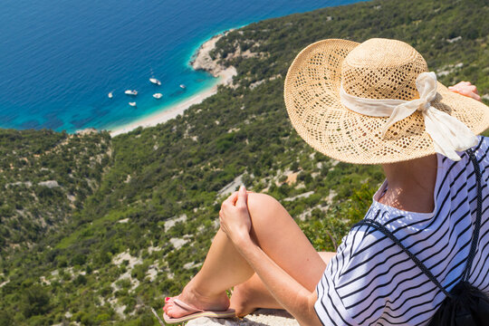 Active sporty woman on summer vacations sitting on old stone wall at Lubenice village, wearing straw hat and beach backpack enjoying beautiful coastal view of Cres island, Croatia.