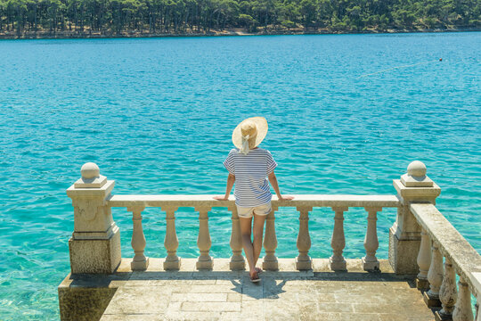 Rear view of woman wearing straw summer hat ,leaning against elegant old stone fence of coastal villa, relaxing while looking at blue Adriatic sea, on Losinj island Croatia.