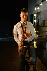 Obraz premium A male musician skillfully plays a brass saxophone, leaning casually against a chair in a dimly lit bar. String lights illuminate the background during a lively evening performance.