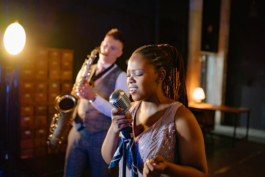 Soulful Black woman sings vintage jazz at an intimate bar performance, accompanied by a saxophonist on stage during an evening concert
