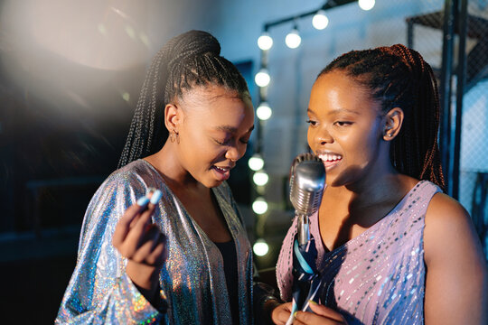 Two Black women in sequined dresses singing into the microphone soul and jazz music together at night on a dark bar stage. The bright stage lights create a warm glow as they sing for the audience