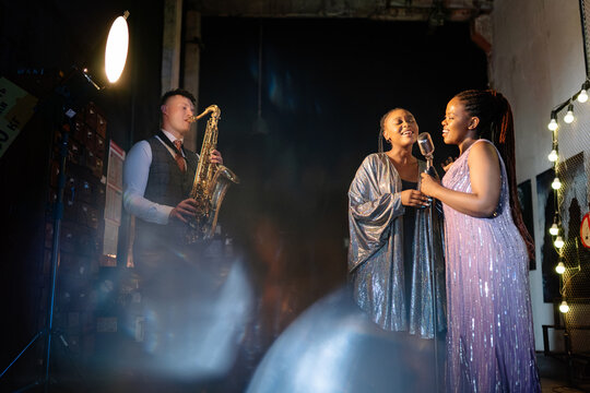 Two soulful black female singers in sequins and a white man saxophonist perform on a dimly lit stage, illuminated by vibrant lens flares during an expressive live session
