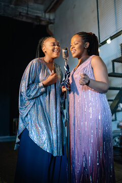 Two Black women dressed in sparkling sequined gowns sing soulful jazz music together into a vintage microphone while performing on a dimly lit bar stage during a night event