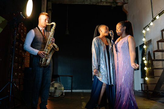 A male saxophonist and two female singers perform live jazz in a stylish, warm-lit urban studio at night