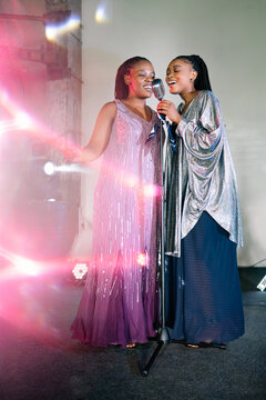 Two graceful Black women in sparkling evening dresses perform soul music together on a dark stage under a bright spotlight while singing into a shared vintage microphone during a lively night concert