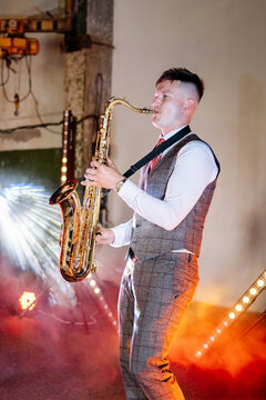 Young man in formal grey suit plays golden tenor saxophone during a vibrant evening party event inside an industrial venue with colorful stage lights and atmospheric smoke effects filling the room