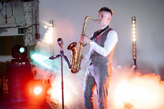 A saxophonist in a grey suit plays a gold saxophone on stage, surrounded by bright theatrical spotlights and atmospheric smoke during a live evening performance
