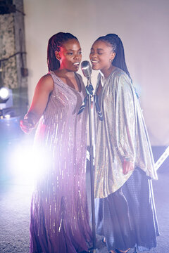 Two talented Black women in sequined dresses sing soulfully into a vintage microphone under bright spotlights on a dimly lit stage