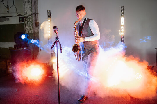 White man musician playing a saxophone on stage during a night performance surrounded by thick fog and bright light beams while wearing formal vest attire for evening event in industrial indoor venue
