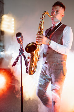 A male musician plays a soulful jazz saxophone solo under a spotlight, surrounded by thick fog and shadows on a formal stage