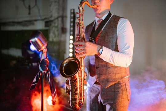 A jazz musician plays a gold saxophone on stage, surrounded by vibrant spotlights, atmospheric smoke, and a vintage microphone during a live evening concert