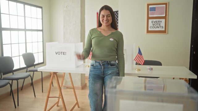 Woman points finger toward camera over transparent ballot box at a polling table inside a building; civic pride.