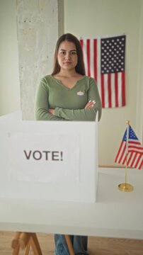 Woman stands with arms crossed at a ballot box wearing a green sweater and 'i voted' sticker beside a small american flag in a building polling booth; civic duty pride.