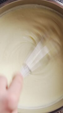 Whisk stirs white sauce smoothly, close up in saucepan. White sauce forms creamy top layer for classic moussaka recipe. White sauce works well for eggplant casserole or Greek baked dish.