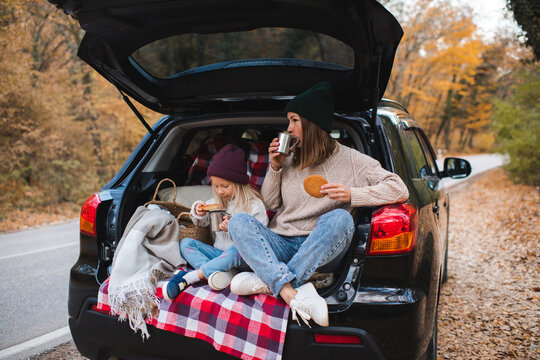 Happy mother with child girl having picnic in car trunk sitting on checkered blanket in woods over autumn leaves. Fall season. Childhood.