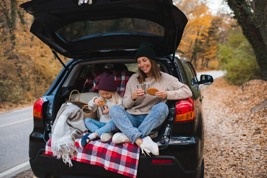 Happy mother with child girl having picnic in car trunk sitting on checkered blanket in woods over autumn leaves. Fall season. Childhood.