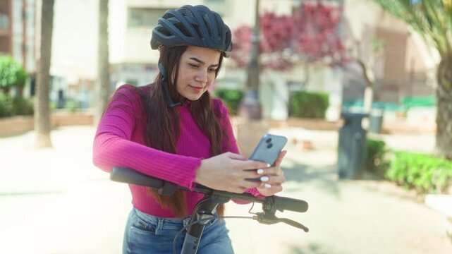 Woman on electric scooter holding smartphone and texting, wearing helmet and pink sweater with hands on handlebar, standing on a city street; serenity focus.