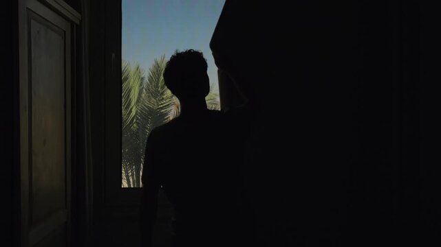 Hot climate lifestyle as man closes blackout curtains to reduce harsh sunlight entering home through window with palm tree outside, improving indoor comfort